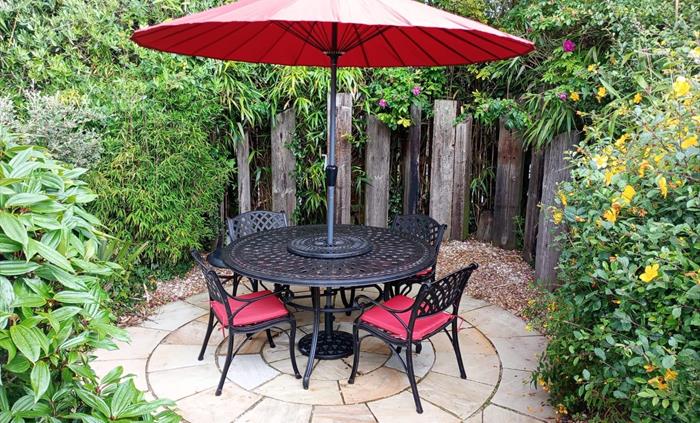 A black iron table and chairs with a lovely red parasol and cushions kept in the garden shed. It sits on a new round patio.  Perfect for summer evenin